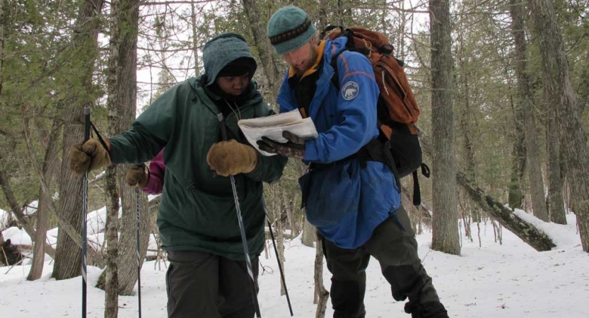 Two people wearing winter gear examine a map in a snowy, wooded area. 